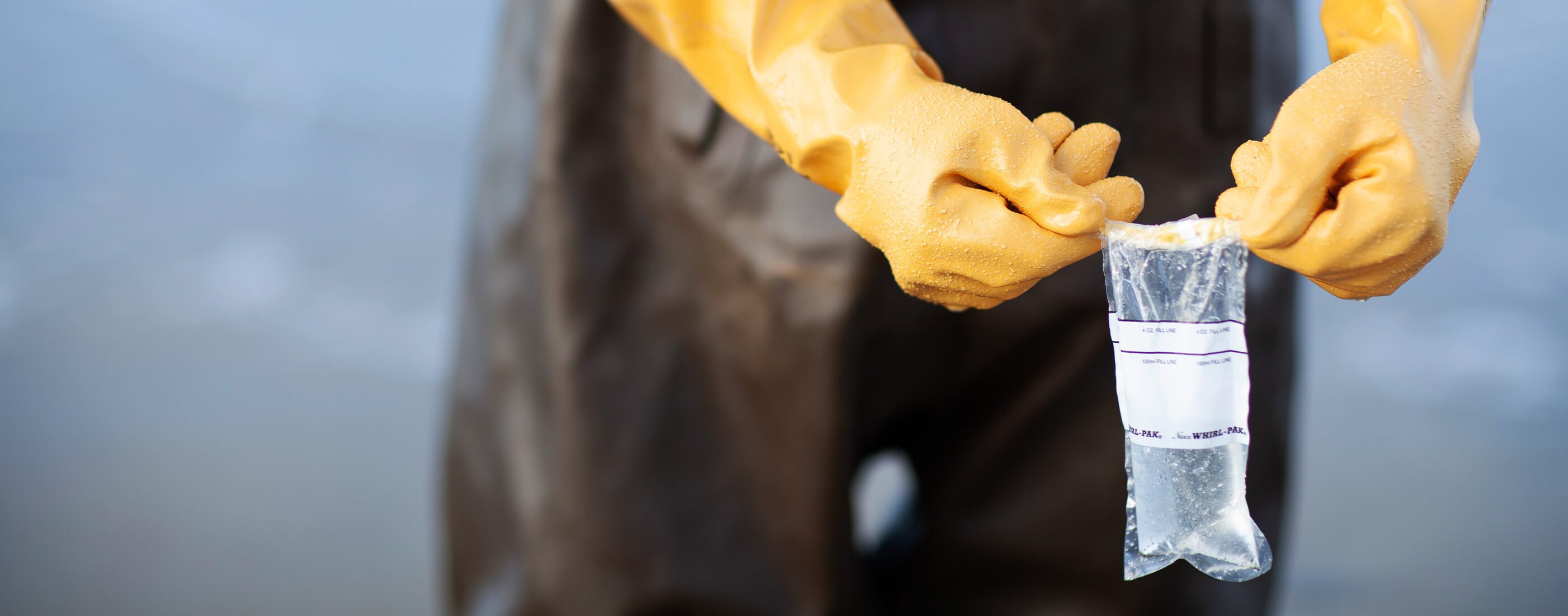 Hands in yellow safety gloves hold up filled water sample bag on beach.